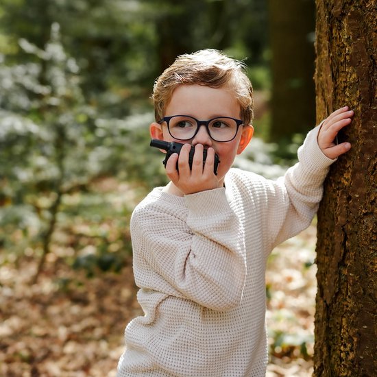 Een jongen in een bos, achter een boom, met een walkie talkie bij zijn mond.