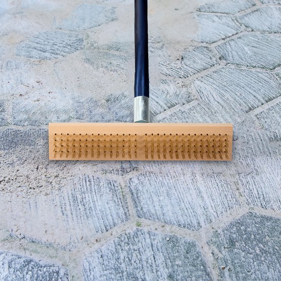 Houten bezem met metalen borstels bovenop een grijze stenen ondergrond.