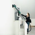 Vrouw met stofmasker gebruikt een lange groene schuurmachine op een muur naast een trap.