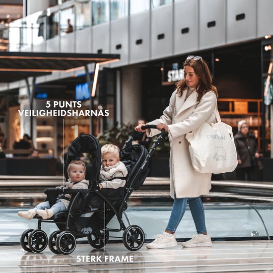 Zwarte dubbele buggy met twee kinderen, een vrouw duwt de buggy in een winkelcentrum.