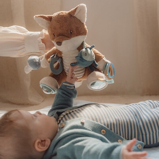 Een baby reikt naar een bruine vos knuffel die door een hand wordt vastgehouden.