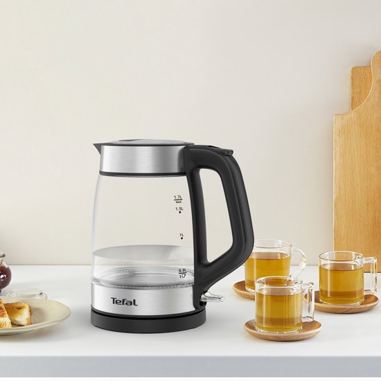 Glazen waterkoker met zilverkleurige en zwarte details, naast theekoppen gevuld met thee.