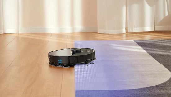 Zwarte robotstofzuiger rijdt van een houten vloer naar een paars en grijs vloerkleed.