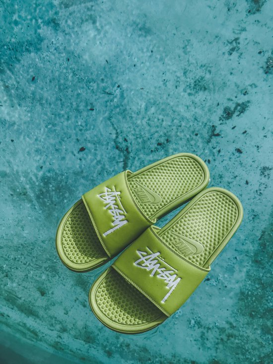 Lichtgroene slippers met witte Stüssy logo's, drijvend in blauw water.