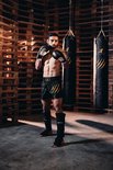 Man met zwarte bokshandschoenen, zwarte vechtsportbroek en scheenbeschermers.