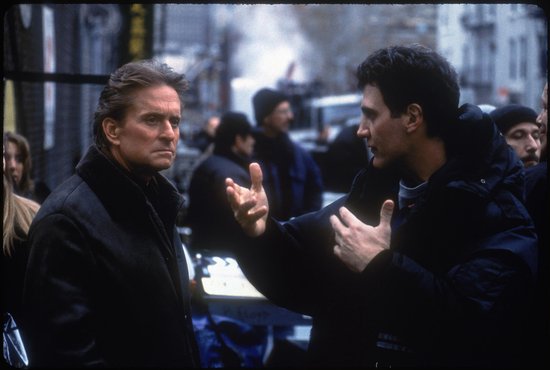 Michael Douglas in een donkerbruine jas met bontkraag, pratend met een man in een donkere jas.