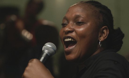 Een vrouw met gevlochten haar zingt luidkeels in een microfoon. Ze draagt een zwart shirt en zilveren oorringen.