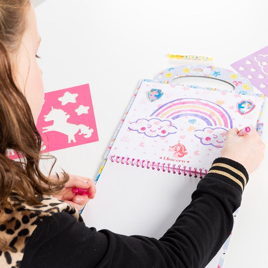 Une fille portant une veste à imprimé léopard dessinant dans un livre d'activités Licorne avec arc-en-ciel et pochoirs.
