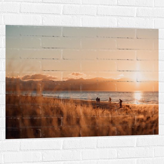 WallClassics - Muursticker - Zon Verdwijnend achter Bergen bij Strand - 100x75 cm Foto op Muursticker