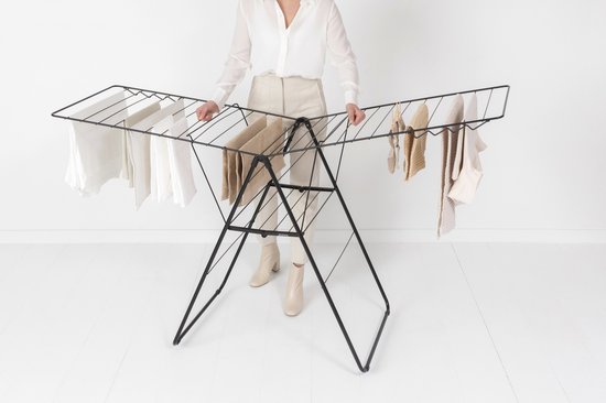 Vrouw in beige kleding staat bij een zwart droogrek met wasgoed.