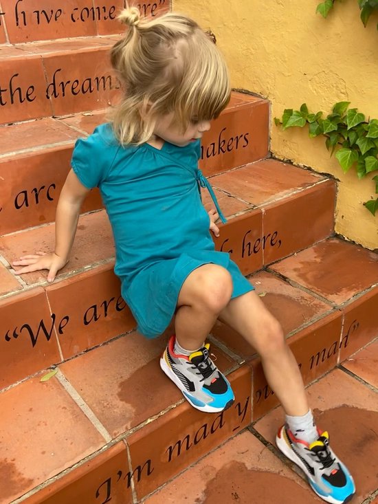 Meisje met blond haar in een turquoise wikkeljurk en gekleurde sneakers zit op een trap.