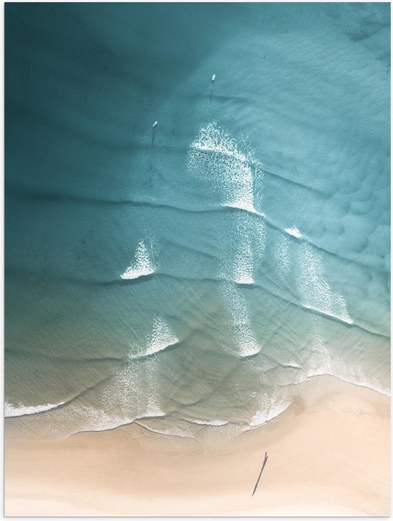 Bovenaanzicht van turquoise oceaangolven die op een zandstrand breken, met twee surfers in het water.