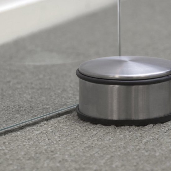 Ronde, metalen deurstopper op een grijze, zachte vloerbedekking, met een glazen deur.