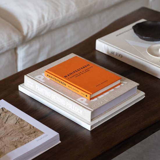 Een stapel boeken op een houten tafel, met de oranje 'Manifesteren' bovenop.