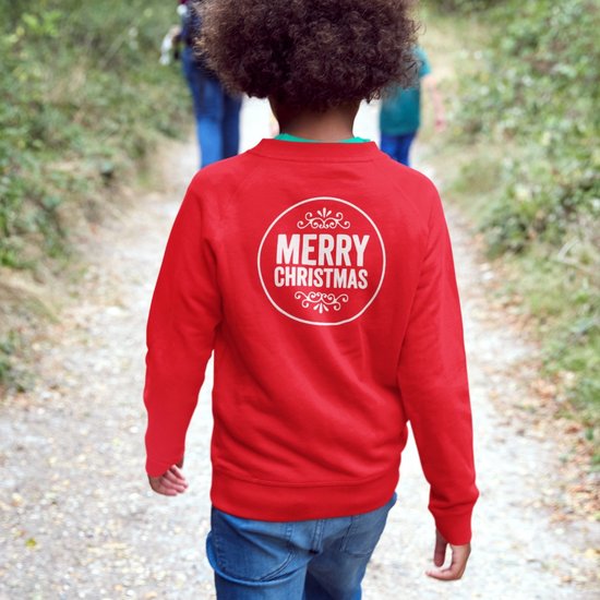 Garçon portant un pull de Noël rouge avec imprimé 'Merry Christmas' dans le dos, debout sur un sentier forestier.