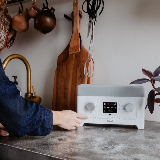 Een hand drukt op de witte Teufel radio met grijs rooster, staand op een betonnen aanrecht.