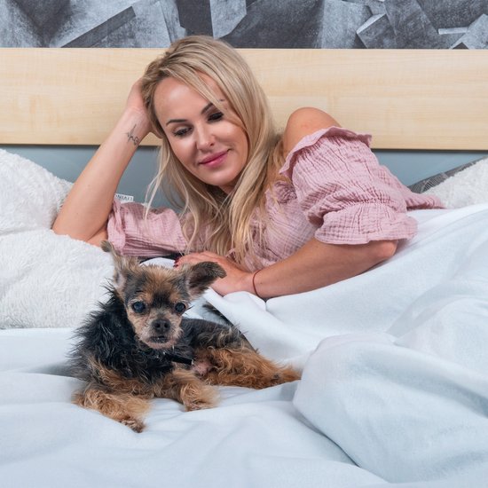 Een vrouw met blond haar en een roze top ligt in bed met een kleine hond.