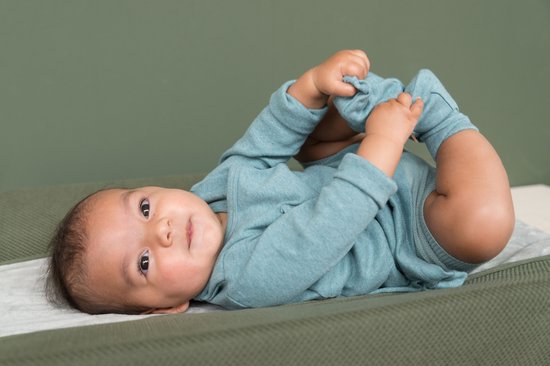 Bébé en body bleu clair est allongé sur le dos, tirant sur ses propres pieds sur une surface verte.