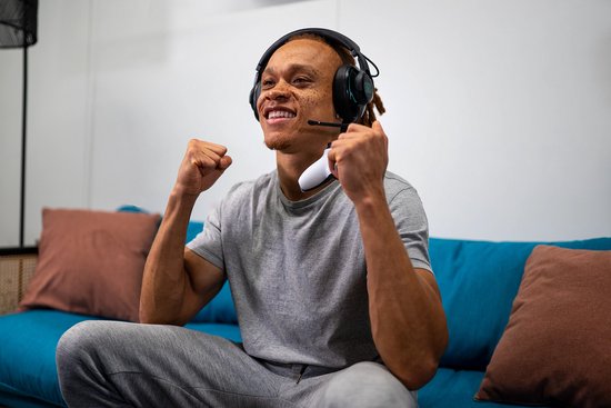 Vrolijke man met een JBL Quantum headset, juichend tijdens het gamen op een bank.