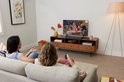 Une barre de son JBL noire est posée sur un meuble TV en bois sous une télévision, deux femmes regardent la télévision depuis un canapé.