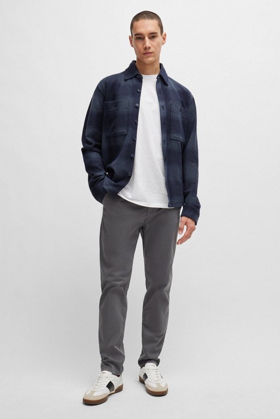 Man in een donkerblauw geruite openvallende jas met lange mouwen, witte t-shirt, grijze chino broek en witte sneakers.