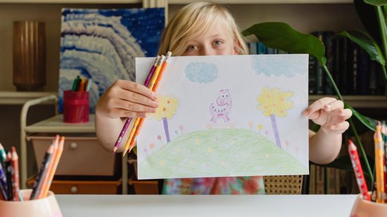 Meisje met blonde haren houdt een kindertekening omhoog, met potloden in haar hand.