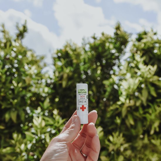 Hand houdt witte SOS stick van INCIA met rode kruis en groene tekst vast tegen een achtergrond van groene bomen en lucht.