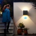 Vrouw loopt 's avonds een trap op bij een verlichte, zwarte kubuswandlamp aan een muur.