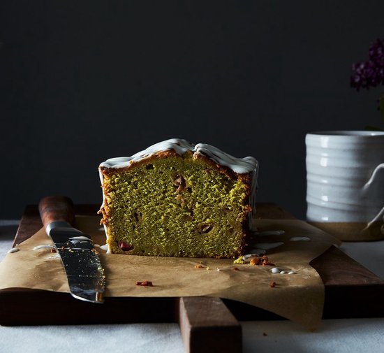 Een stuk groene matcha cake met witte glazuur, geserveerd op bruin bakpapier op een houten plank, met een mes ernaast.