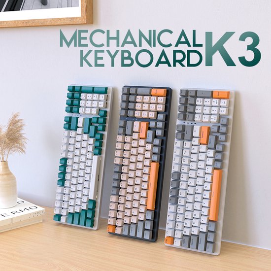 Drie mechanische toetsenborden K3 op een bureau met een plant en boeken, in de kleuren groen/wit, oranje/grijs en oranje/grijs.