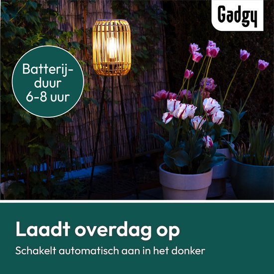 Brandende rotan lantaarn op driepoot in de tuin bij schemering. Laadt overdag op, brandt 6-8 uur.