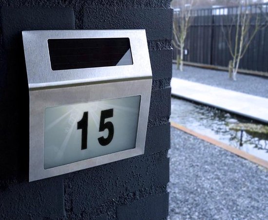 Zilverkleurige huisnummerlamp met nummer '15' verlicht, naast een vijver en met bomen op de achtergrond.