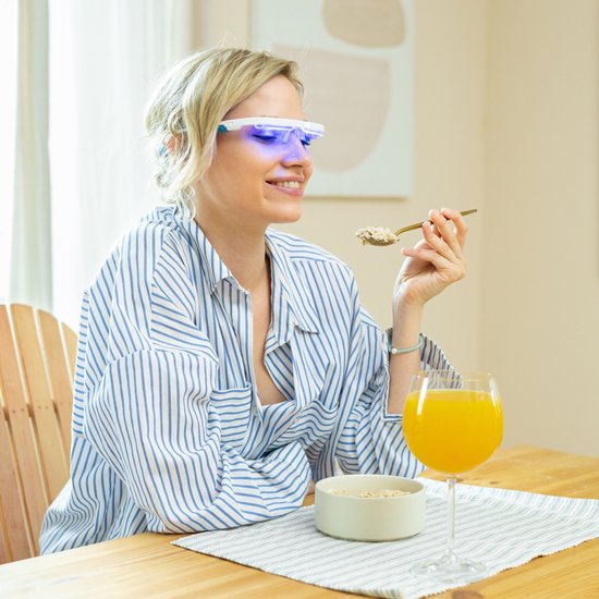 Vrouw met blauwe lichtbril eet ontbijt aan tafel, gestreepte blouse.