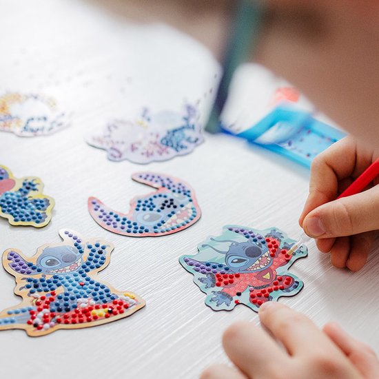 Mains d'enfant réalisant un magnet Stitch diamond painting avec des perles rouges et bleues. Stitch porte un costume rouge, avec un stylo.