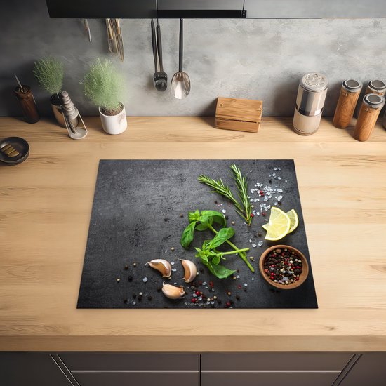 Zwarte inductiebeschermer met betonprint, bedekt met kruiden, knoflook, citroen en peperkorrels, op een houten aanrechtblad.