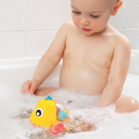 Een baby zit in bad en speelt met een gele badeend visje met rode vinnen en blauwe staart.