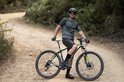 Man met een mountainbike op een bosweg, draagt een donkergroen shirt en korte broek, helm en bril.