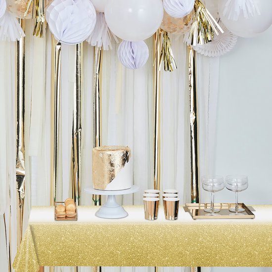 Een feesttafel met een gouden glitter tafelkleed, witte en gouden slingers en ballonnen, een witte taart met gouden decoratie en macarons.