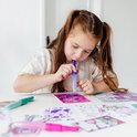 Meisje met bruine staartjes gebruikt een paarse spray pen op een roze Gabby's Poppenhuis sjabloon met figuren en glitter-effecten.
