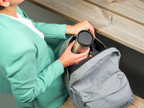 Vrouw in een groen pak legt een roestvrijstalen beker met grijs deksel in een grijze rugzak.