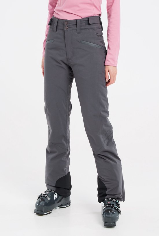 Femme portant un col roulé rose et un pantalon de ski gris, avec des chaussures de ski.