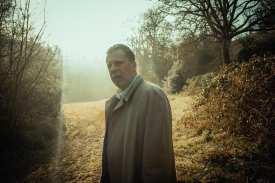 Een oudere man met grijs haar, staand in een mistig bos in een beige trenchcoat.