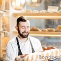 Bakker met baard draagt een zwart schort en houdt een lang, versgebakken brood vast.