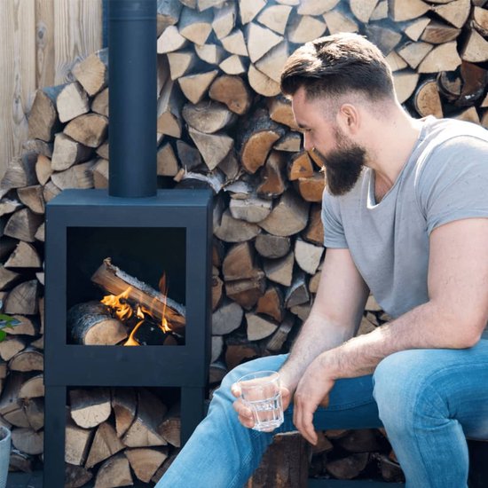 Man kijkt naar brandende zwarte buitenkachel, met stapels hout op de achtergrond.