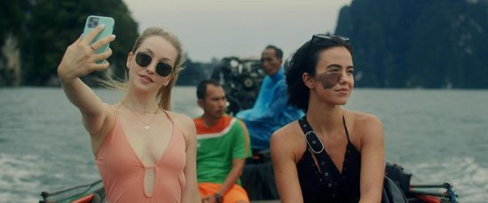 Twee jonge vrouwen, een met blond haar en zonnebril in een roze badpak, de ander met donker haar en een zwart badpak, maken een selfie op een boot. Op de achtergrond varen mannen.