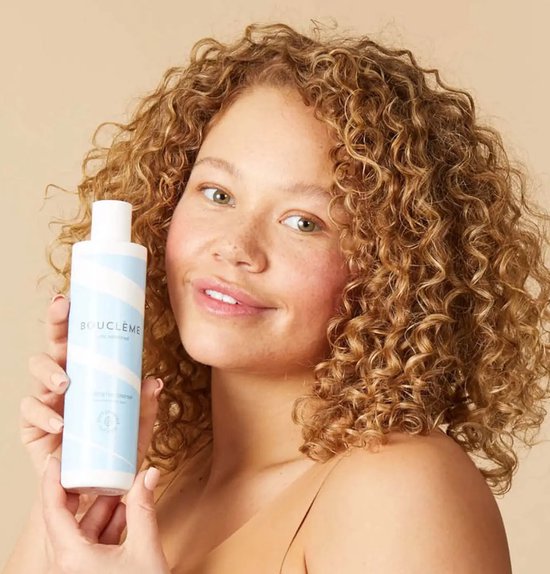 Vrouw met krullend haar houdt een fles bouclème hydraterende haarreiniger vast. Fles heeft blauwe en witte strepen.