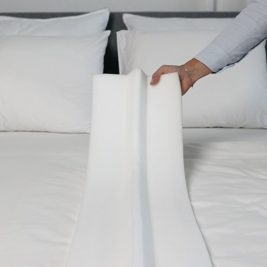 Hand houdt een witte, schuimrubberen matraswig omhoog boven een bed met witte lakens.