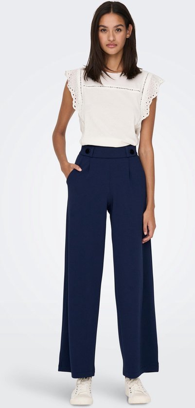 Model in donkerblauwe wijde broek met hoge taille, knopen, steekzakken en witte top met kanten details.