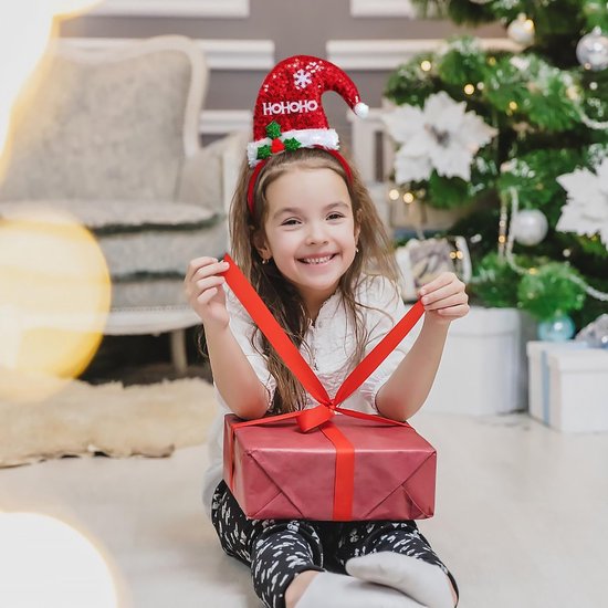 Een lachend meisje met een kerstmuts met 'HO HO HO' en glitters, zittend voor een kerstboom met een cadeau.