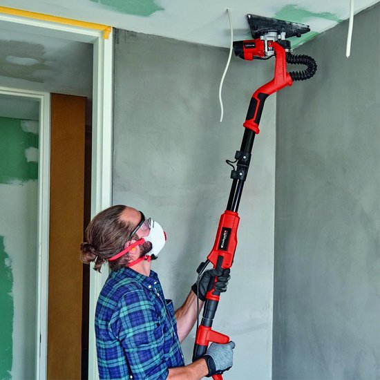 Man met veiligheidsbril en masker schuurt een plafond met een lange rode schuurmachine.